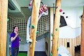 Kind beim Klettern an einer Holzwand im Indoor-Spielbereich des Familienhotels Amiamo im Salzburger Land, beaufsichtigt von einer lächelnden Betreuerin, die ein aktives und kinderfreundliches Hotelkonzept hervorhebt.