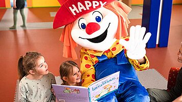 Clown Happy liest zwei Mädchen ein Buch vor