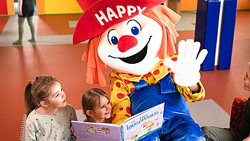 Clown Happy liest zwei Mädchen ein Buch vor