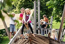 Kinder spielen gemeinsam mit einer Kinderbetreuerin auf einem Outdoor Spielplatz des Familienhotels Allgäuer Berhof.