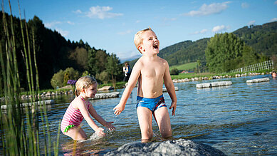 Zwei fröhliche Kinder spielen im flachen Wasser eines Naturbades mit Blick auf grüne Hügel im Familienhotel Schreinerhof im Bayerischen Wald.