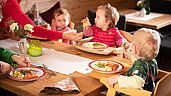 Mutter sitzt mit vier Kleinkindern am Tisch im familienfreundlichen Restaurant des Familienhotels Landgut Furtherwirt in Tirol. Die Kinder sitzen im Hochstuhl und essen Fischstäbchen mit Kartoffeln vom bunten Kinderteller.