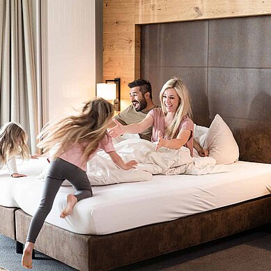 Familienhotels Hotelzimmer mit viel Platz für Spaß: Paar sitzt im Hotelbett, die beiden Töchter stürmen lachend zu den Eltern.