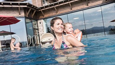 Eine Mutter ist mit ihrer kleinen Tochter, die Schwimmflügel trägt, im Pool. Die Sonne scheint, beide sind glücklich. So schön ist es im Familienhotel Allgäuer Berghof.