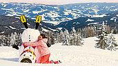 Ein Kind umarmt einen umgefallenen Schneeman in Kaernten. Der Ausblick im Winter kann sich sehen lassen und zeigt ein tolles Panorama.