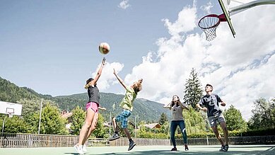 Familienhotel Alpenhof Dolomit Family. Teens spielen gemeinsam auf dem Sportplatz Basketball.