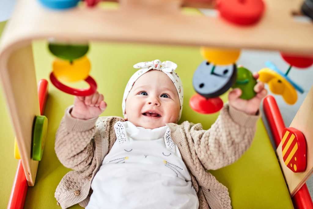Glückliches Baby spielt mit einem Spielebogen im Babyspielbereich des Familienhotels Aigo welcome family in Oberösterreich.