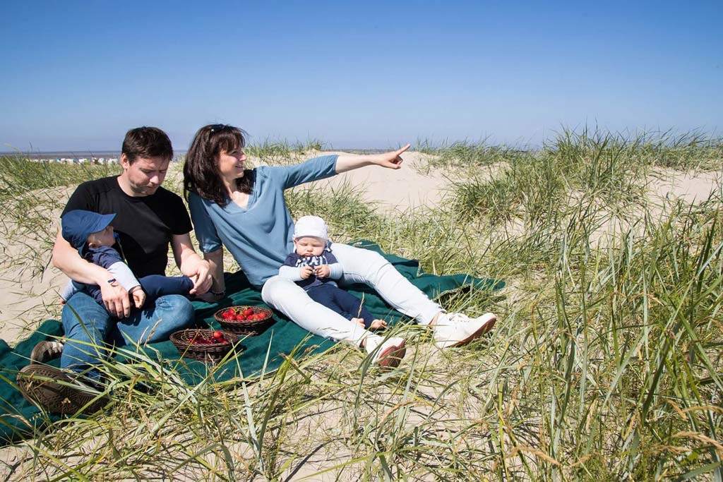 Eine Familie genießt ein Picknick auf einer grünen Decke in den Dünen mit Blick auf die Nordsee, wobei die Mutter auf etwas in der Ferne deutet, während der Vater und zwei kleine Kinder sich entspannen und Erdbeeren essen, was die familienfreundliche Umgebung und die Nähe des Familienhotels Deichkrone zum Strand hervorhebt.