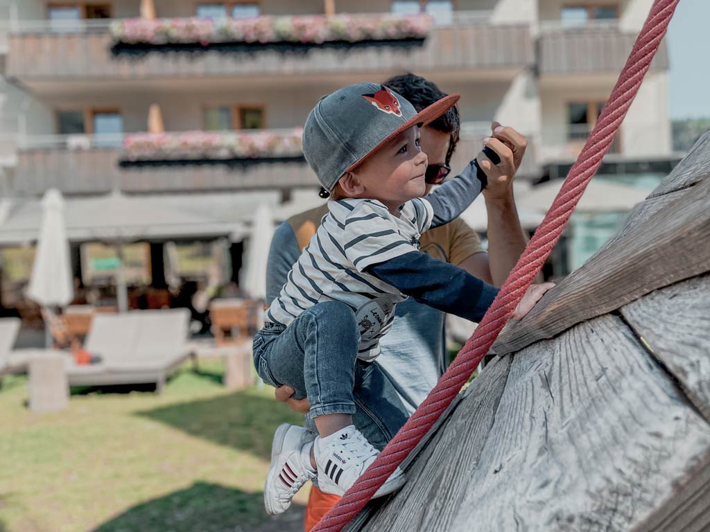 Ein Vater hält sein Baby fest, während dieses versucht auf dem Outdoor-Spielplatz eine Kletterwand hinauf zu klettern im Familienhotel Gorfion in Liechtenstein.
