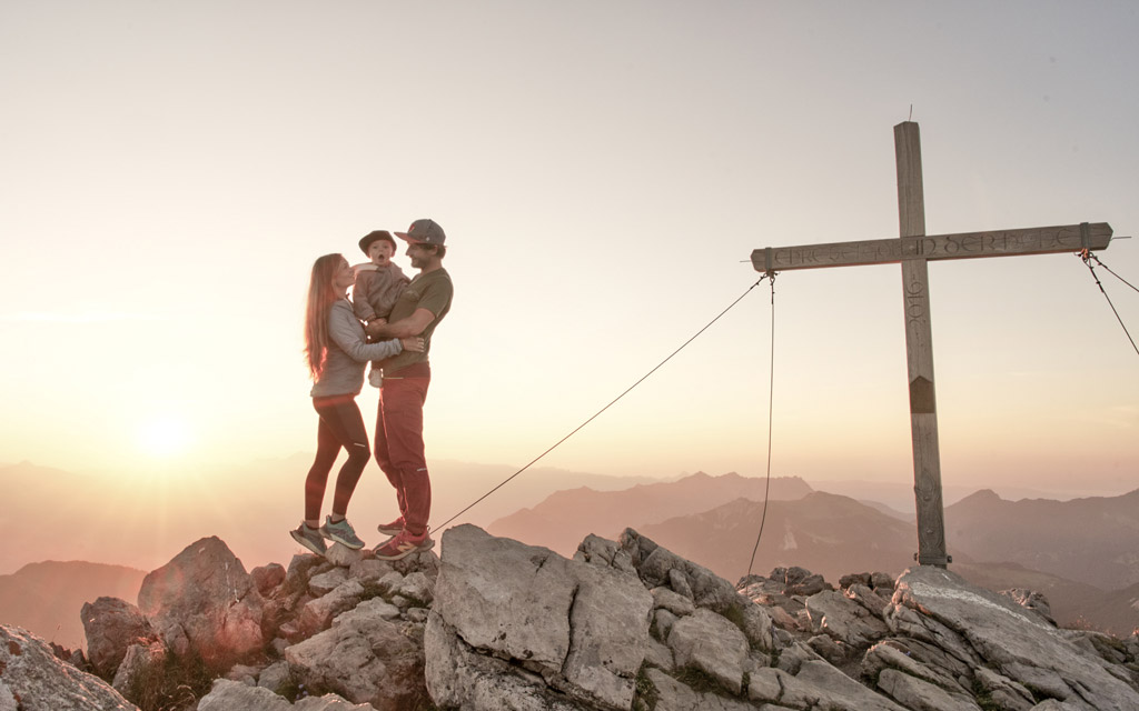 Eltern genießen mit ihrem Kind auf dem Arm den Sonnenaufgang am Gipfel in Liechtenstein