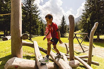 Ein Junge geht über ein großes Holzspielzeuggerüst auf dem Outdoorspielplatz am Almfamilyhotel Scherer in Tirol 