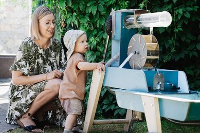 Mutter beobachtet ihr Kleinkind beim Spielen am Wasserspielplatz im Garten des Familienhotels Tirolerhof.