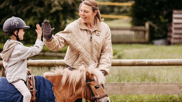 Ein Kind mit Reithelm sitzt auf einem kleinen Pony und gibt einer lächelnden Trainerin am Zaun ein High-Five; das Pony trägt eine blaue Decke, im Hintergrund eine grüne Koppel.