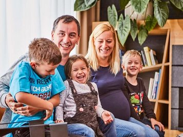 Familie mit drei Kindern sitzt zusammen auf einer gemütlichen Couch in der Lobby des Familienhotels Aigo im Böhmerwald.