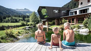 Am hauseigenen Badesee mit Steg sitzen Kinder in der Sonne. Freut euch auf perfekte Urlaubsmomente im Familienurlaub im Alphotel Tyrol in Südtirol.