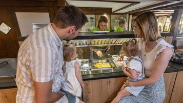 Ein Mann und eine Frau halten jeweils ein Kind im Arm und schauen einer Hotel-Mitarbeiterin beim Zubereiten eines Eier-Omletts zu. 