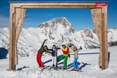 Eine Familie posiert fröhlich auf Skiern vor einem Holzrahmen mit Bergpanorama im Skigebiet nahe dem Familienhotel Ramsi in Kärnten