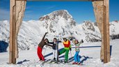 Eine Familie posiert fröhlich auf Skiern vor einem Holzrahmen mit Bergpanorama im Skigebiet nahe dem Familienhotel Ramsi in Kärnten