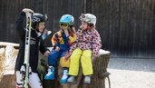 Kinder sitzen mit Ski-Ausrüstung auf einem Holzstamm und freuen sich auf die Piste im Familienhotel Bavaria im Allgäu.