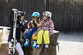 Kinder sitzen mit Ski-Ausrüstung auf einem Holzstamm und freuen sich auf die Piste im Familienhotel Bavaria im Allgäu.