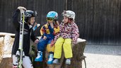 Kinder sitzen mit Ski-Ausrüstung auf einem Holzstamm und freuen sich auf die Piste im Familienhotel Bavaria im Allgäu.