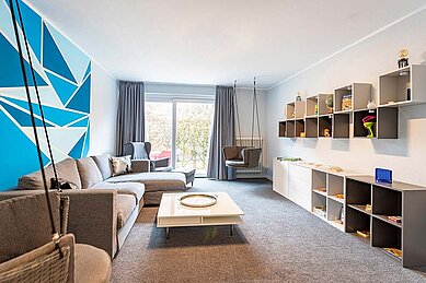 Moderne und gemütliche Jugendlounge im Familienhotel Deichkrone mit einer stylishen blauen Grafikwand, komfortablen Sofas, einer Hängeschaukel und Regalen voller Spiele, perfekt für Teenager zum Entspannen und Treffen mit neuen Freunden.