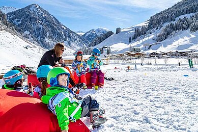 Familie macht eine Pause vom Skifahren und sitzt im Schnee bei Sonnenschein.