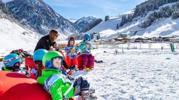 Familie macht eine Pause vom Skifahren und sitzt im Schnee bei Sonnenschein.