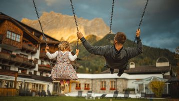 Vater und Kind schaukeln im Hotelgarten vor leuchtender Bergkulisse bei dramatischem Himmel.
