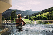 Eine Frau steht im Infinitypool und genießt die Aussicht auf die umliegenden Berge im Familienhotel Post Family Resort im Salzburger Land.