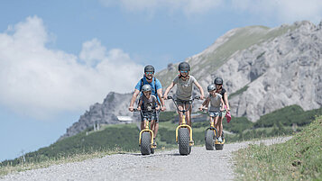 Eltern sind mit ihren Kindern auf dem Naturspielplatz und die Tochter schaukelt auf der Schaukel im Familienhotel Liechtenstein.