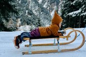 Kind liegt lachend auf einem Holzschlitten im Schnee beim Winterurlaub im Familienhotel Kindl mit Bergpanorama im Hintergrund.