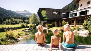 Drei Kinder genießen die Sonne am Naturteich des Familienhotel Alpura Retreat in Südtirol.
