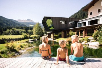Drei Kinder genießen die Sonne am Naturteich des Familienhotel Alpura Retreat in Südtirol.