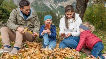 Eine Familie sitzt an einem Herbsttag in der Natur zwischen bunten Blaettern und genießt gemeinsam einen Moment im Freien vor einer Bergkulisse.