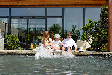Drei Kinder sitzen am Beckenrand des Pools des Familienhotels Ulrichshof und lassen ihre Füße ins Wasser hängen.