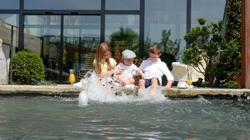 Drei Kinder sitzen am Beckenrand des Pools des Familienhotels Ulrichshof und lassen ihre Füße ins Wasser hängen.