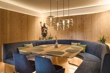 Stilvoller Essbereich im Zauchenseehof mit runder Polsterbank, großem Holztisch, modernen Glaspendelleuchten und dekorativer Statue an der Wand.