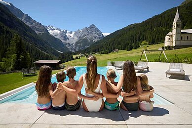 Kinder und Mama sitzen nebeneinander am Beckenrand und genießen die Aussicht auf die Berge im Familienhotel Bella Vista in Südtirol.