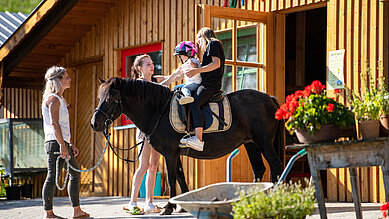 Zwei Kinder sitzen vor der Reithalle des Familienhotels Der Böhmerwald auf einem Pferd. Die Mutter führt das Pferd mit den beiden Kindern.