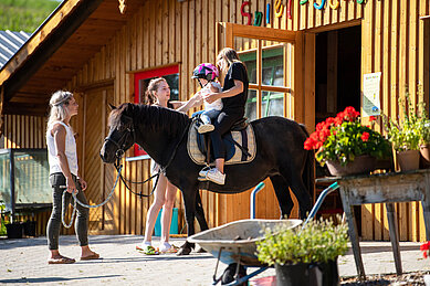 Zwei Kinder sitzen vor der Reithalle des Familienhotels Der Böhmerwald auf einem Pferd. Die Mutter führt das Pferd mit den beiden Kindern.