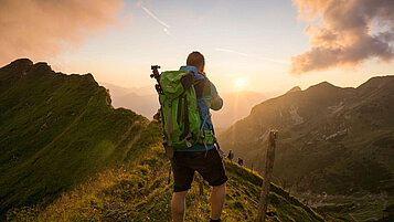Ein Mann beim Wandern steht auf dem Gipfel eines Berges im Allgäu und genießt die Aussicht und den Sonnenuntergang. Die Berge im Hintergrund rahmen den Mann ein.
