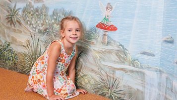 Ein Kind sitzt vor einer bemalten Wand mit Feenmotiv in der Magischen Abenteuerwelt des Familienhotels Kaiserhof in Tirol, aufgenommen im Bereich Feengrotte.