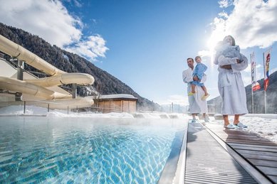 Familie im Bademantel mit zwei Kleinkindern am Rand eines dampfenden Outdoor-Thermalpools im Schnee, Bergkulisse und Wasserrutsche im Hintergrund.