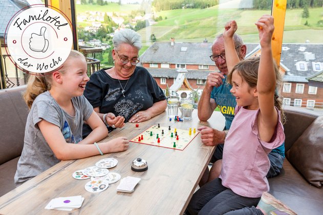 Familie spielt ein Brettspiel am Tisch auf einer Terrasse; ein Kind jubelt mit erhobenen Armen, im Hintergrund Blick auf ein Dorf in grünen Hügeln, Badge „Familotel der Woche“.