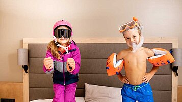 Kinder posieren auf Hotelbett im Skianzug und Badebekleidung für das Angebot vom Familienhotel Das Bayrischzell