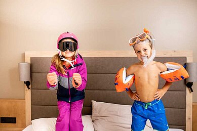 Kinder posieren auf Hotelbett im Skianzug und Badebekleidung für das Angebot vom Familienhotel Das Bayrischzell