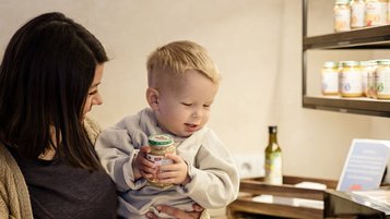 Eine Mutter hält ihr Baby im Arm, der ein Gläschen mit Babybrei in der Hand hält