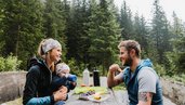 Eltern mit Kleinkind sitzen an einem Picknicktisch im Wald und genießen eine Pause vom Wandern.