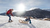 Eine Mutter zieht im Winter einen Schlitten, auf dem zwei kleine Kinder sitzen direkt hinterm Almfamilyhotel Scherer in Tirol 